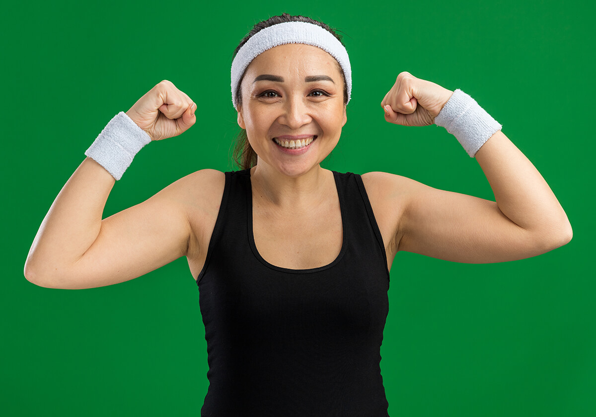 happy young fitness woman with headband armbands smiling confident raising fists standing green wall